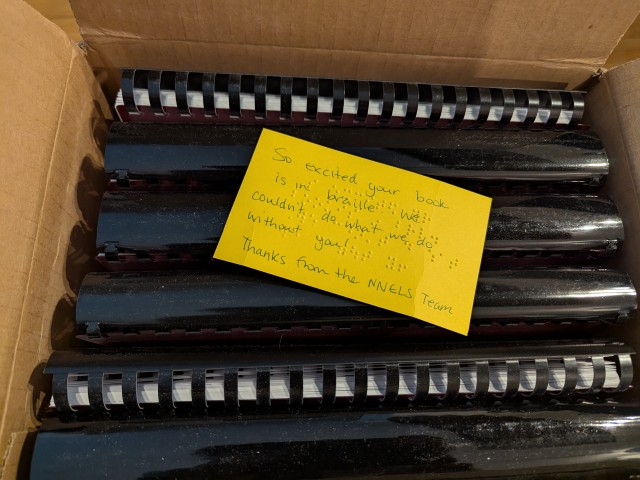 The open box with a yellow note that reads: "So excited your book is in braille. We couldn't do what we do without you! Thanks from the NNELS Team."
